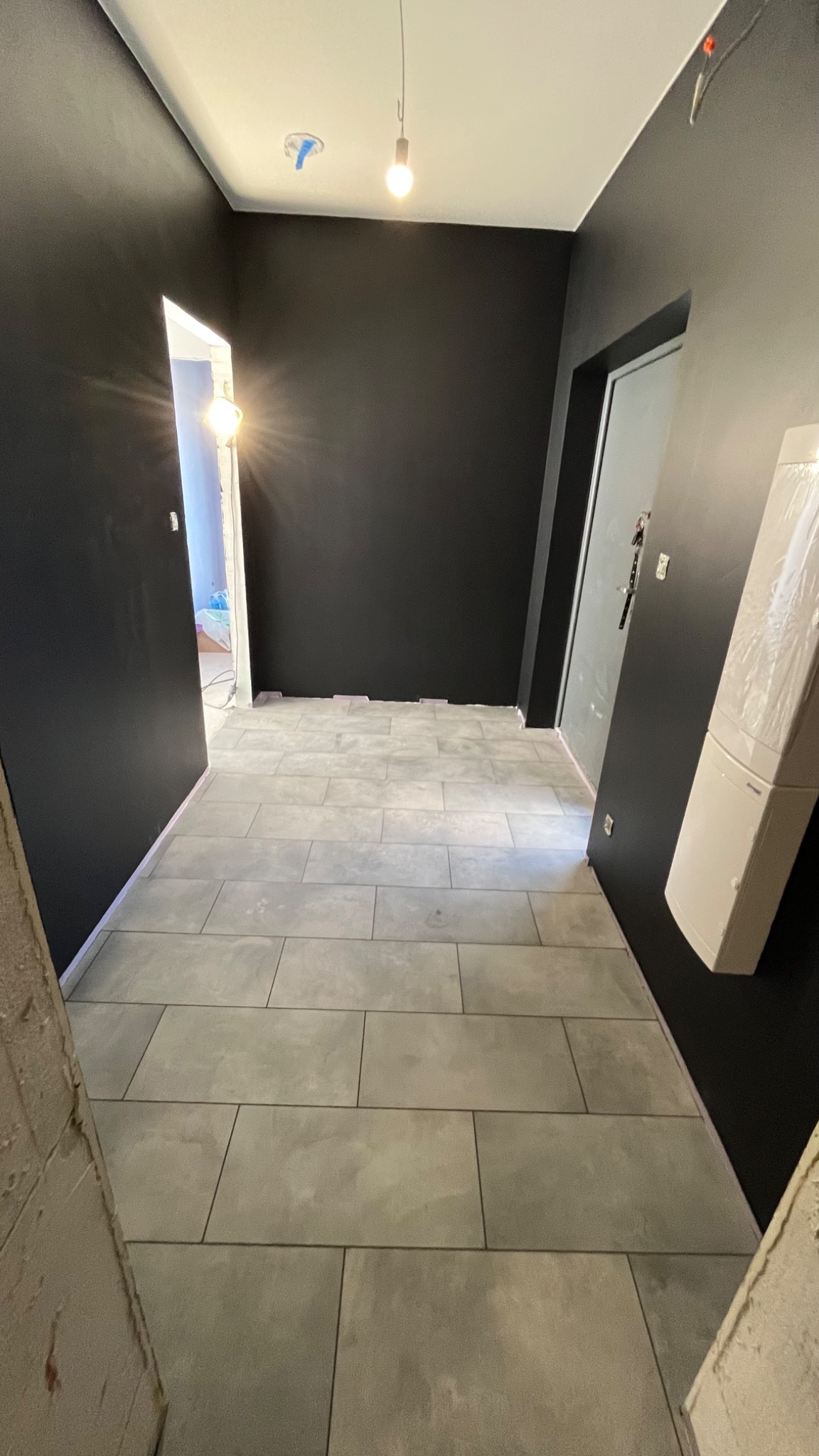 Wykończony korytarz z szarymi płytkami podłogowymi i czarnymi ścianami. Widoczne drzwi i wisząca żarówka. Minimalistyczny design.