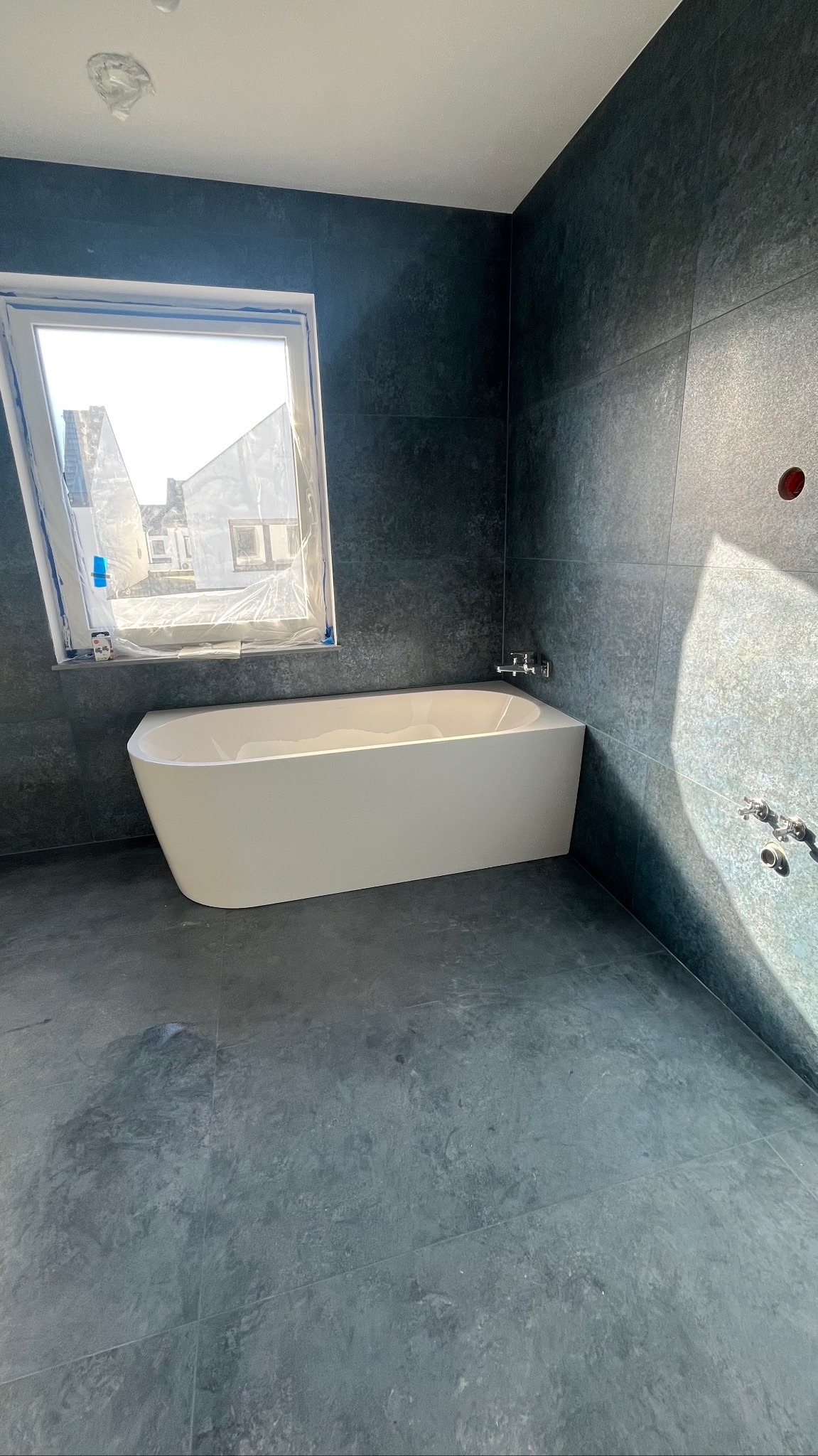 Nowoczesna łazienka z wolnostojącą wanną na tle grafitowych płytek. Okno zabezpieczone folią malarską. Minimalistyczny design, surowy klimat.