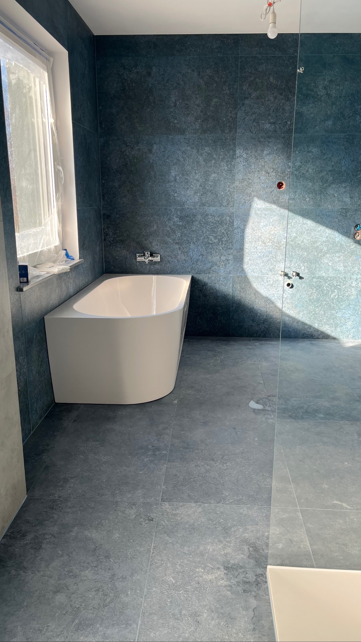 Nowoczesna łazienka z wanną wolnostojącą i szklaną kabiną prysznicową. Ciemne płytki imitujące kamień na ścianach i podłodze. Okno z folią ochronną.