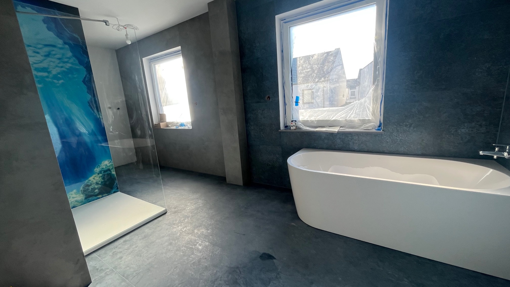 Nowoczesna łazienka w trakcie wykańczania: kabina prysznicowa z motywem morskim, wolnostojąca wanna i okna zabezpieczone folią malarską. Szare płytki na ścianach i podłodze.