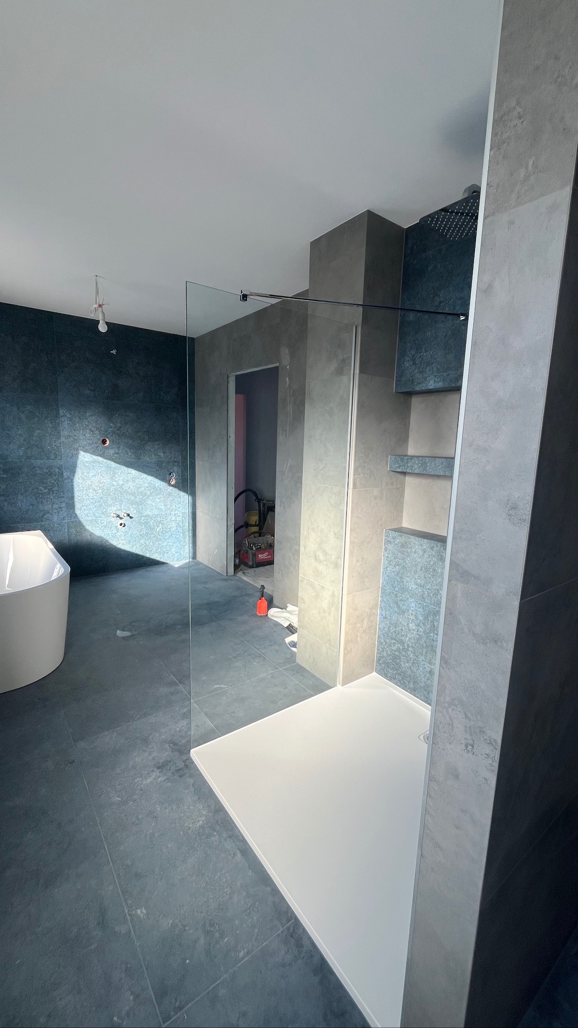 Nowoczesna łazienka w trakcie wykańczania: szare płytki, biała wanna i brodzik, szklana kabina prysznicowa. Widoczne narzędzia i elementy instalacji.