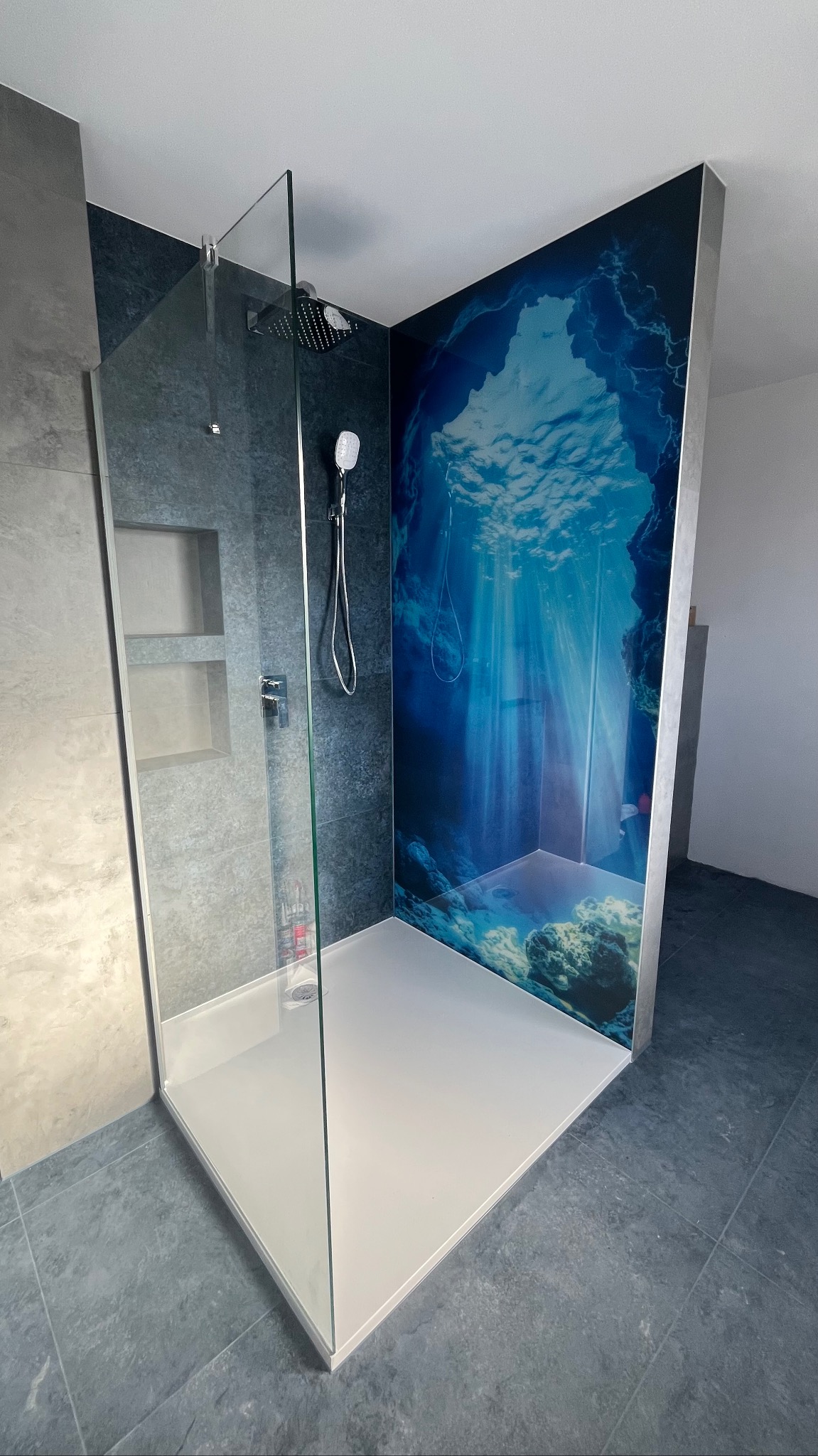 Nowoczesna kabina prysznicowa walk-in z szarą glazurą i panelem z motywem podwodnego świata. Minimalistyczny design, oświetlenie imitujące promienie słoneczne.