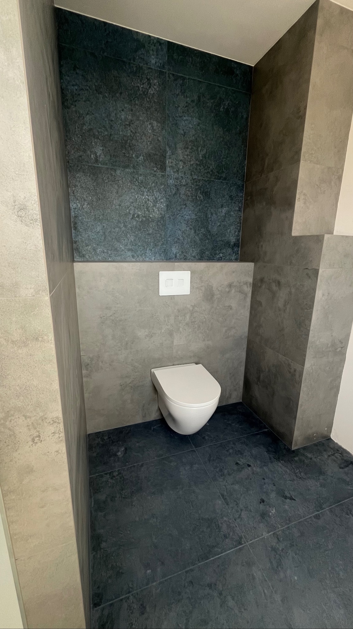 Nowoczesna toaleta podwieszana w łazience wykończonej płytkami w odcieniach szarości i granatu. Minimalistyczny design, widoczny fragment ściany i podłogi.