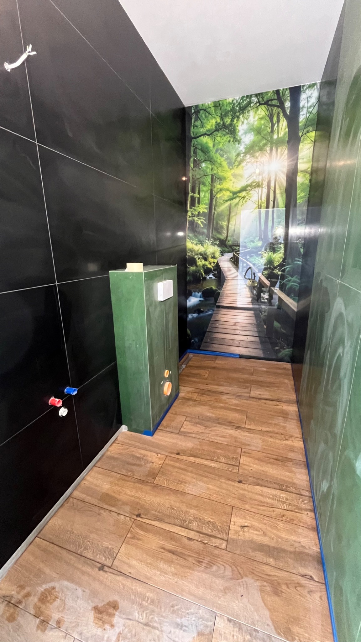 Nowoczesna łazienka w trakcie wykończenia: czarne płytki, drewnopodobna podłoga, zielony stelaż WC i fototapeta z motywem leśnym. Widoczne detale instalacji.