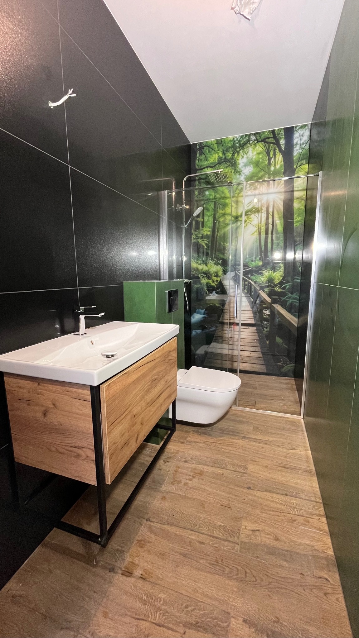 Nowoczesna łazienka z drewnianą szafką pod umywalką, czarnymi płytkami i kabiną prysznicową z motywem leśnym. Uwagę przykuwa nietypowy, zielony kolor zabudowy WC.