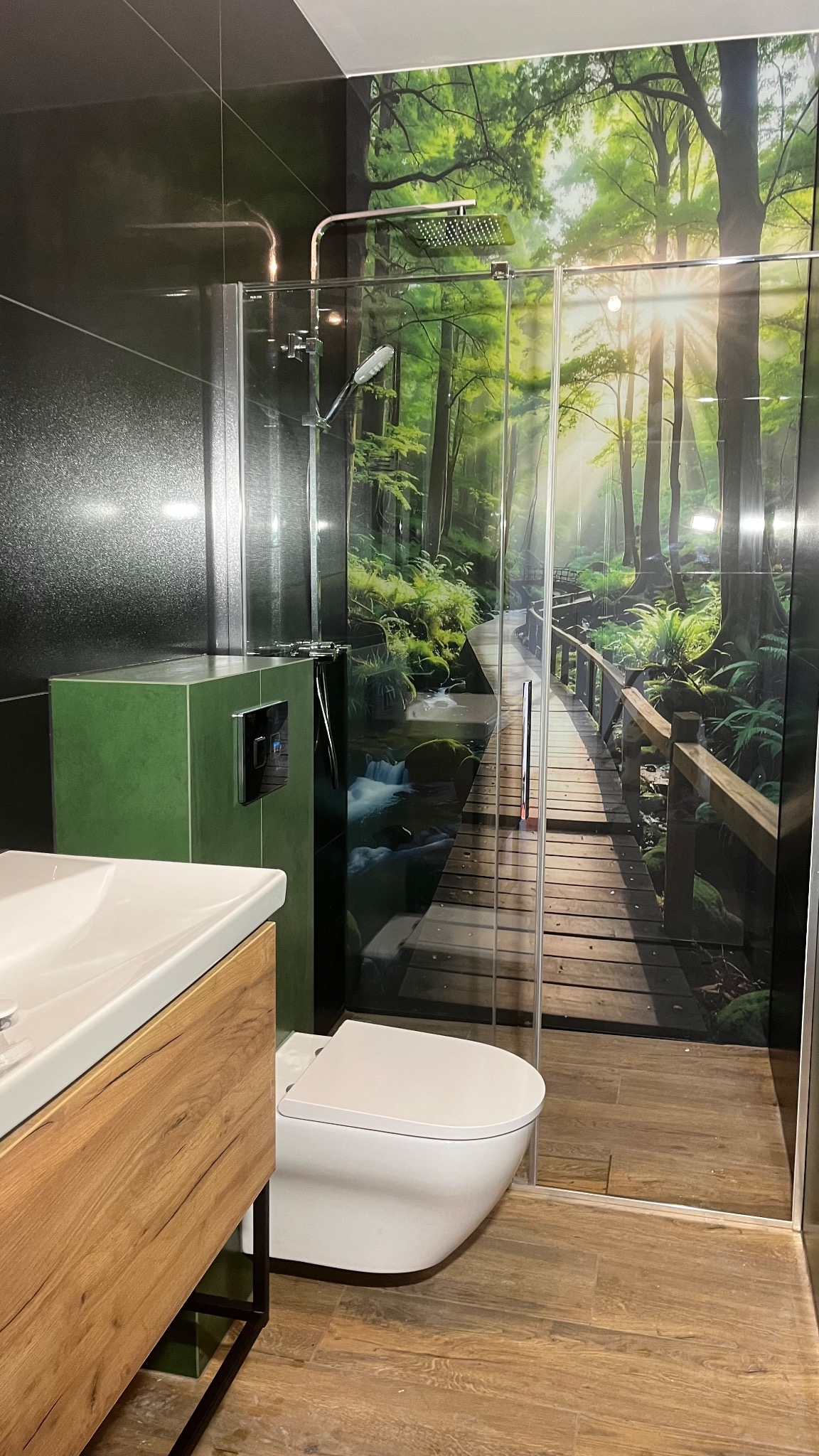 Nowoczesna łazienka z kabiną prysznicową i fototapetą leśną. Umywalka z drewnianą szafką, podwieszana toaleta. Czarna ściana z teksturą i zielony element zabudowy.