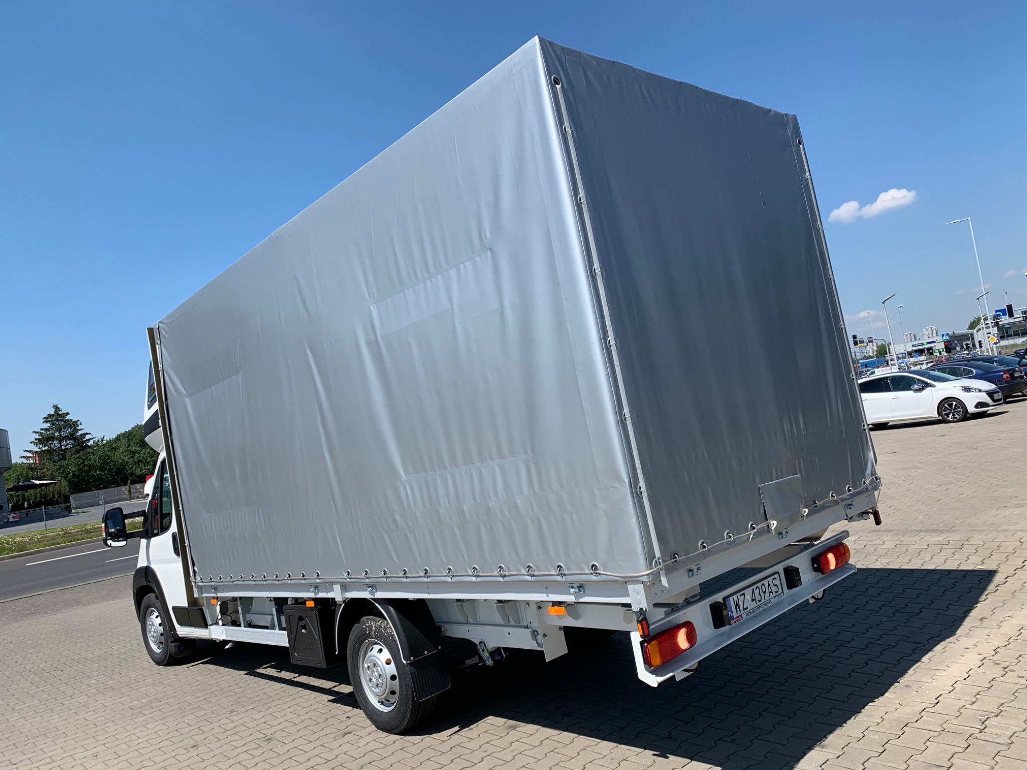 Biały samochód dostawczy z szarą plandeką, zaparkowany na kostce brukowej w słoneczny dzień. Widoczny numer rejestracyjny WZ 439AS.