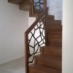 Drewniane schody z nowoczesną balustradą o geometrycznym, czarnym wzorze, widoczne z dołu, na jasnym tle ściany i szarej podłogi.