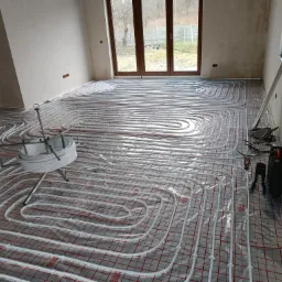 Rozłożona instalacja ogrzewania podłogowego na folii z siatką, w surowym stanie wykończenia, widoczne narzędzia i zwój rury na stojaku.