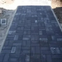 Ułożona, grafitowa kostka brukowa tworząca ścieżkę prowadzącą do furtki. Widoczne obrzeża ścieżki i fragmenty zieleni po bokach.