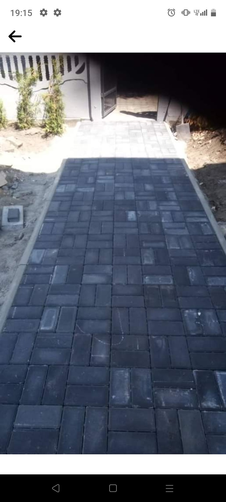 Ułożona, grafitowa kostka brukowa tworząca ścieżkę prowadzącą do furtki. Widoczne obrzeża ścieżki i fragmenty zieleni po bokach.