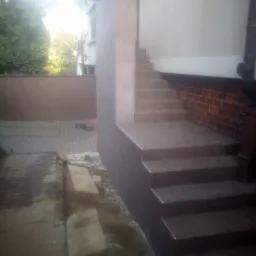 Zewnętrzne schody wejściowe wykończone brązowymi płytkami, z widocznym fragmentem elewacji budynku i chodnikiem w trakcie prac ziemnych.