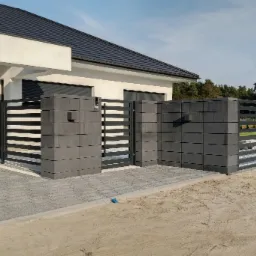 Nowoczesne ogrodzenie z paneli i betonowych słupków w kolorze grafitowym, z przesuwną bramą wjazdową i furtką, na tle domu z ciemną dachówką i brukowanym podjazdem.