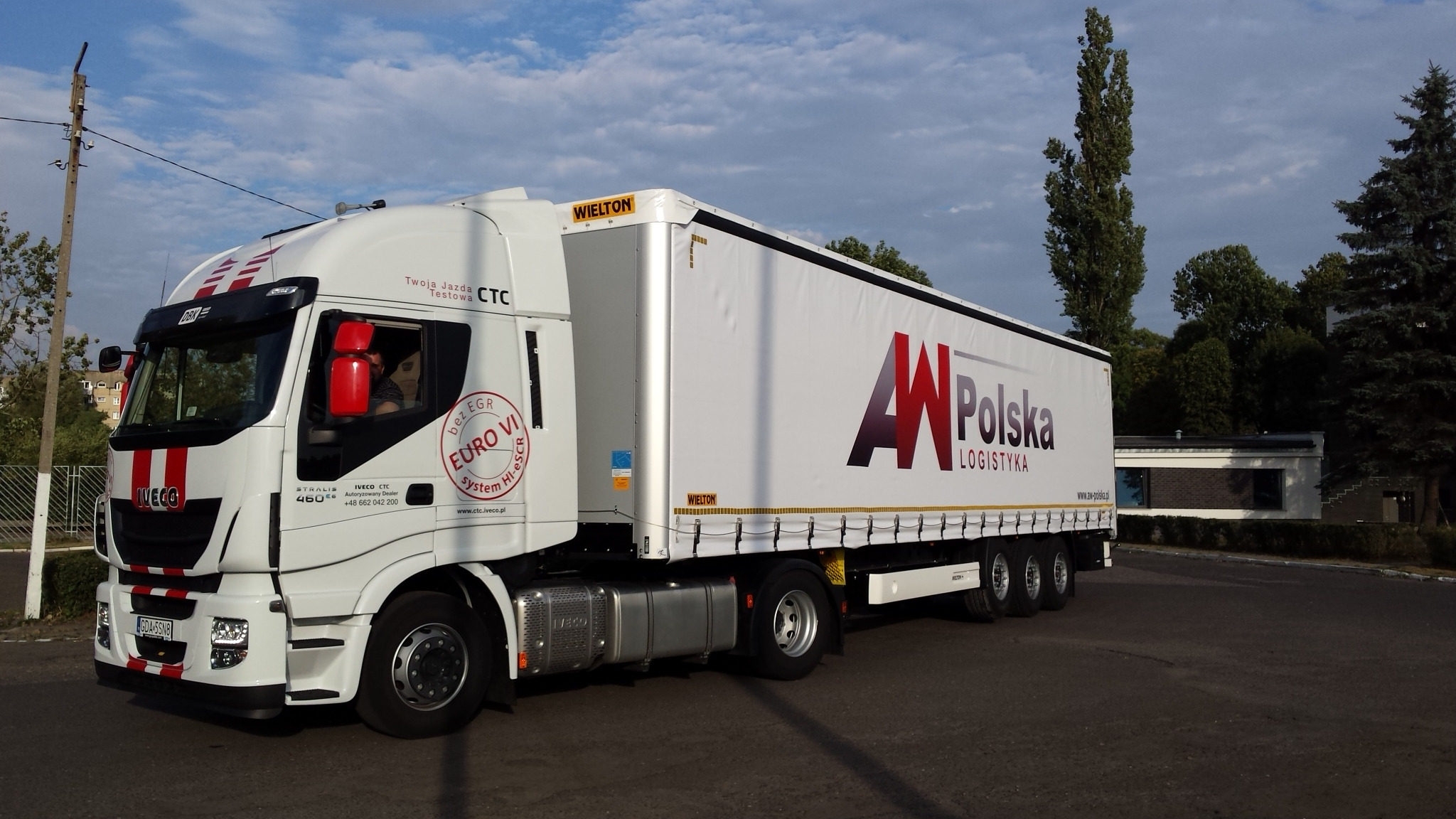 Biała ciężarówka Iveco z naczepą z logo 'Polska Logistyka' na parkingu, widoczne tablice rejestracyjne i kierowca w kabinie.