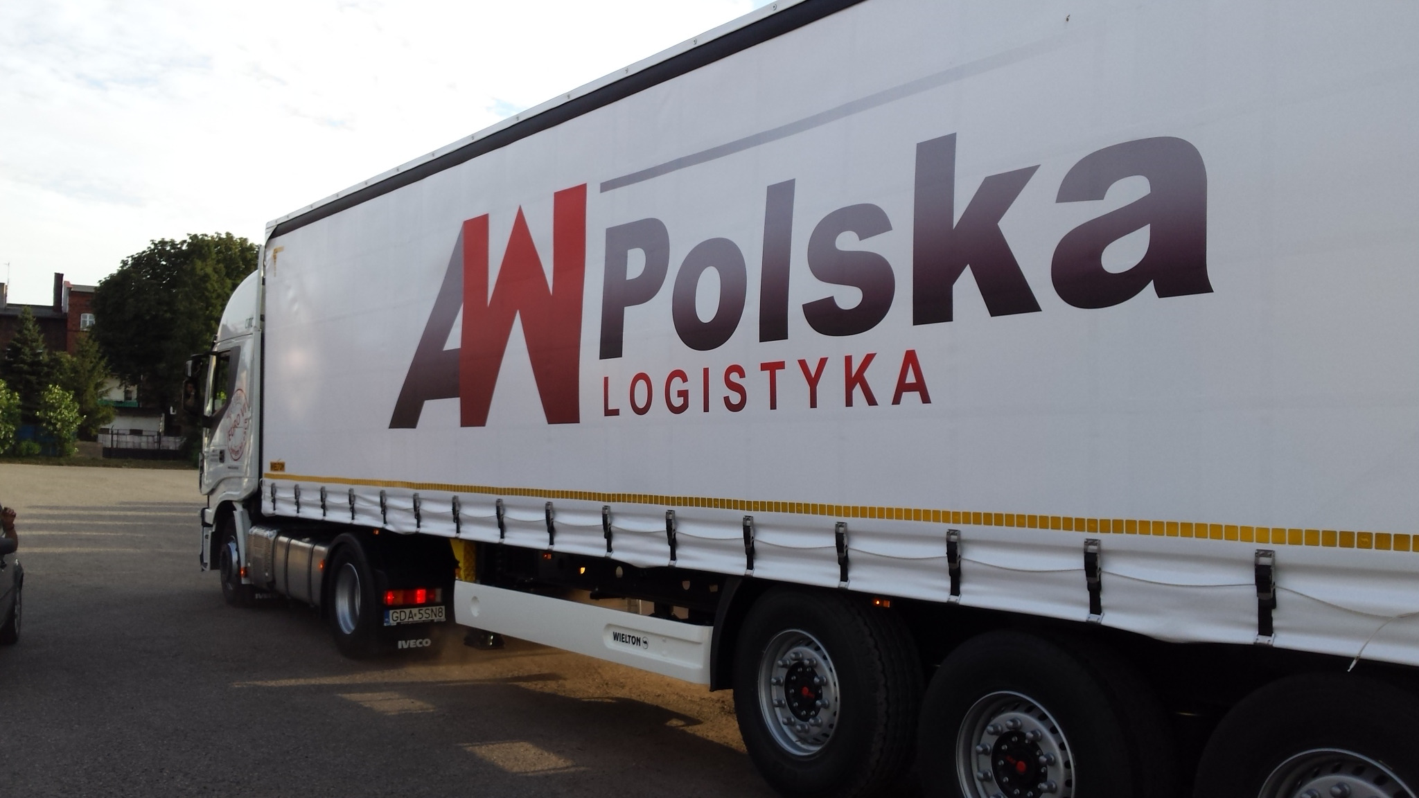 Ciężarówka z białą naczepą, na której widnieje logo firmy 'AW Polska Logistyka', zaparkowana na placu. Naczepa z plandeką zabezpieczoną żółtymi pasami. Widoczny fragment kabiny kierowcy i tablica...