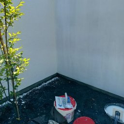Elektroinstal - Świeżo otynkowana ściana zewnętrzna domu z widocznym młodym drzewkiem, wiaderkami z zaprawą tynkarską i narzędziami, zabezpieczona taśmą malarską na górze.