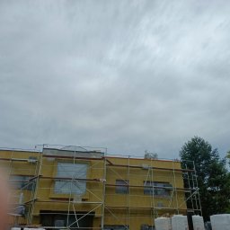 Elektroinstal - Budynek w trakcie ocieplania elewacji, widoczne rusztowania, styropian i worki z materiałami budowlanymi, pochmurne niebo w tle.
