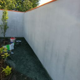 Elektroinstal - Świeżo otynkowana, jasnoszara elewacja budynku z pomarańczowym zabezpieczeniem na górze, widoczne wiadro farby, narzędzia i małe drzewko.