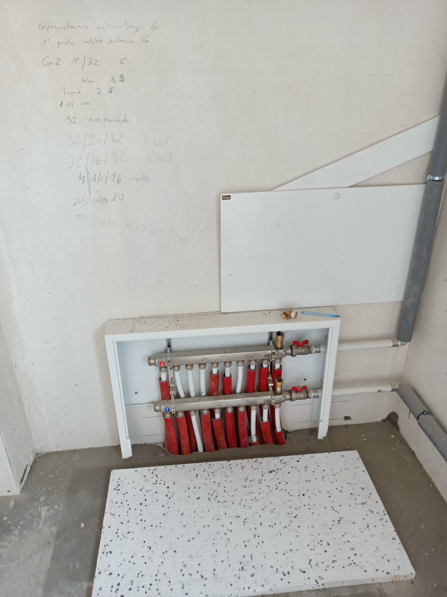 Rozdzielacz ogrzewania podłogowego w białej obudowie, widoczne rury grzewcze w kolorze czerwonym i białym, na ścianie notatki techniczne, na podłodze płyta styropianowa, widok z dołu.