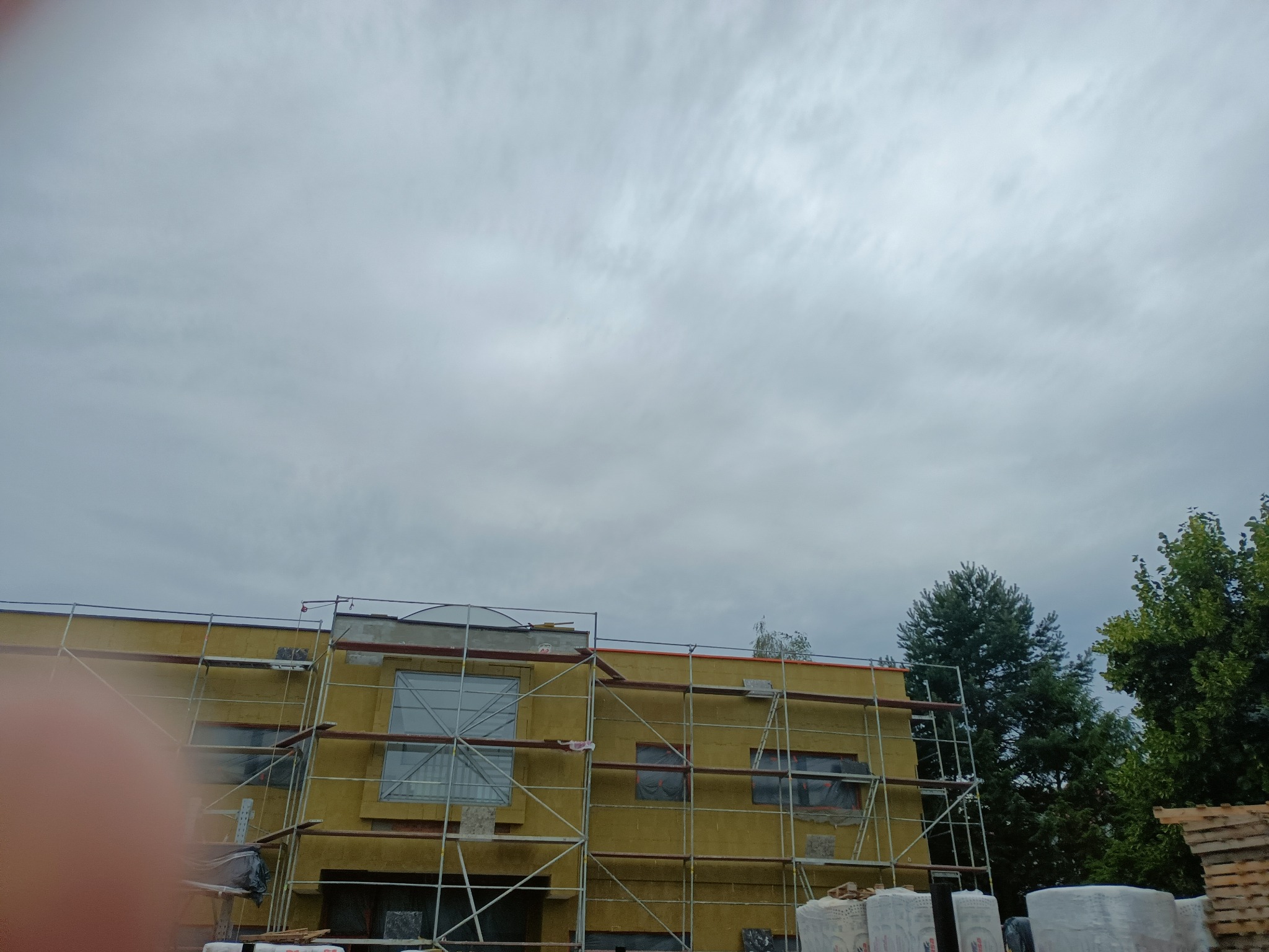 Budynek w trakcie ocieplania elewacji, widoczne rusztowania, styropian i worki z materiałami budowlanymi, pochmurne niebo w tle.