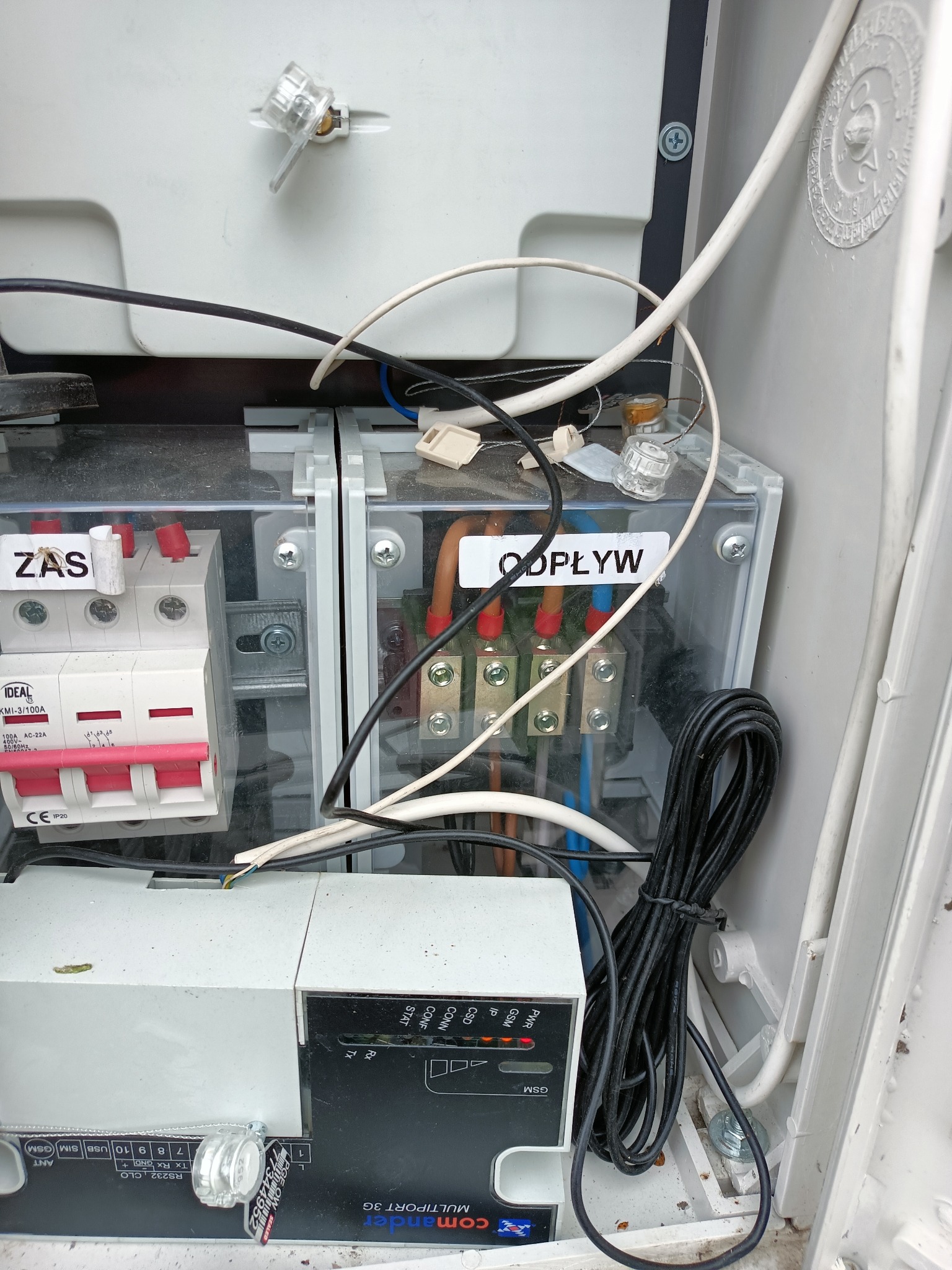 Otwarta skrzynka elektryczna z bezpiecznikami, przewodami i modułem sterującym Comander Multiport 3G. Widoczne oznaczenia 'ZAS' i 'ODPŁYW' na elementach wewnątrz skrzynki.
