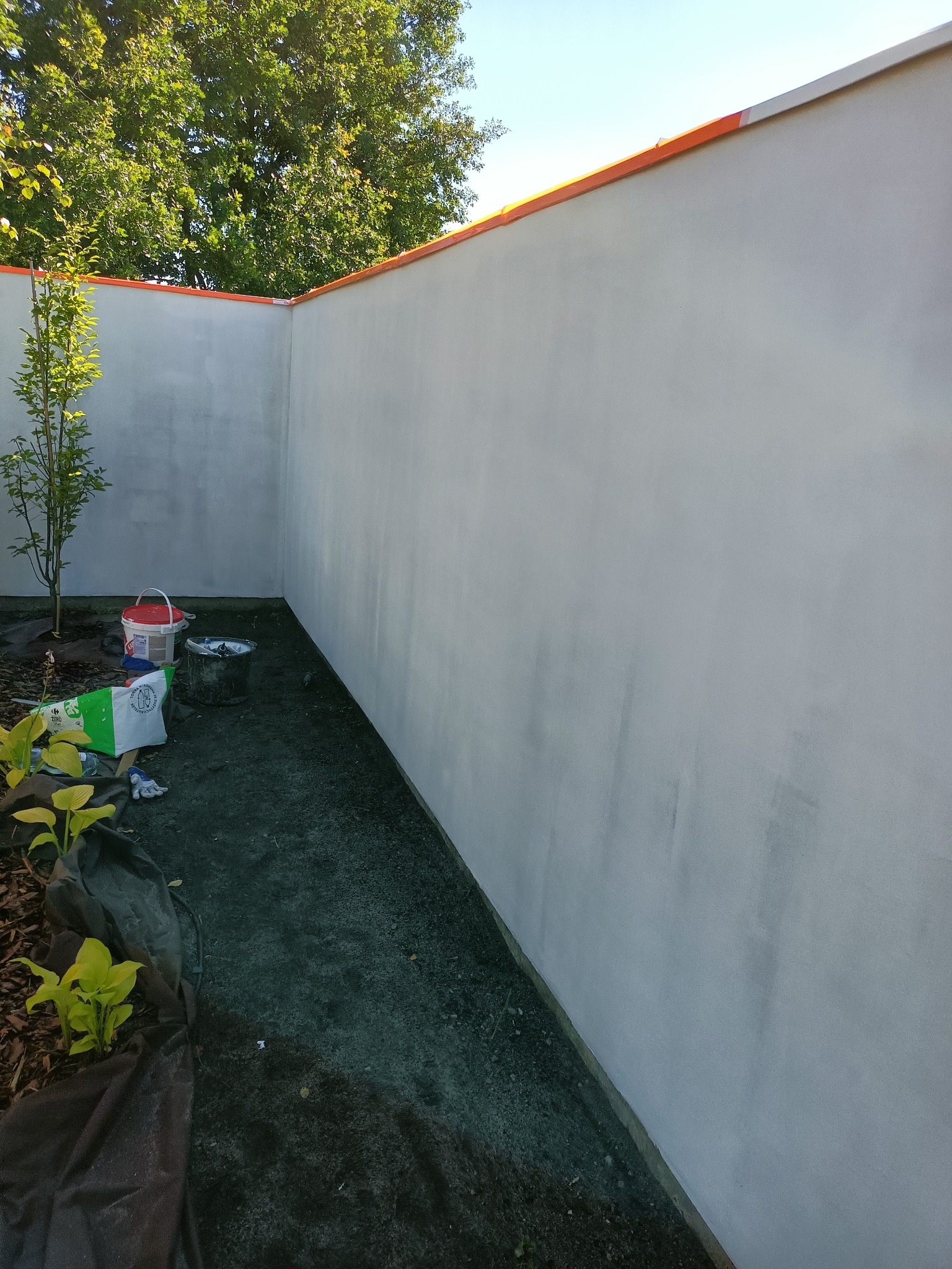 Świeżo otynkowana, jasnoszara elewacja budynku z pomarańczowym zabezpieczeniem na górze, widoczne wiadro farby, narzędzia i małe drzewko.