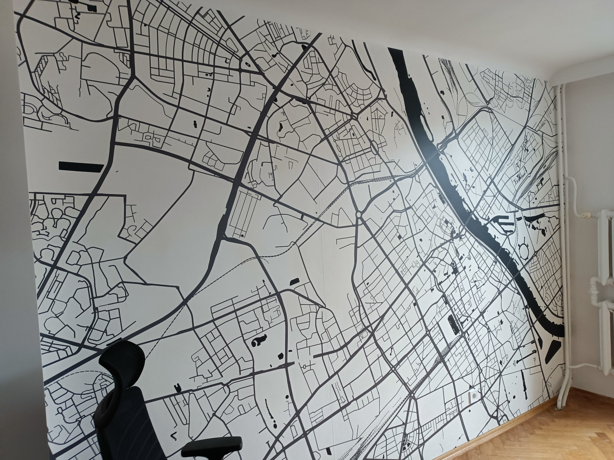 Ściana z tapetą przedstawiającą mapę miasta w czarno-białej kolorystyce, widoczny fragment biurka i grzejnika.