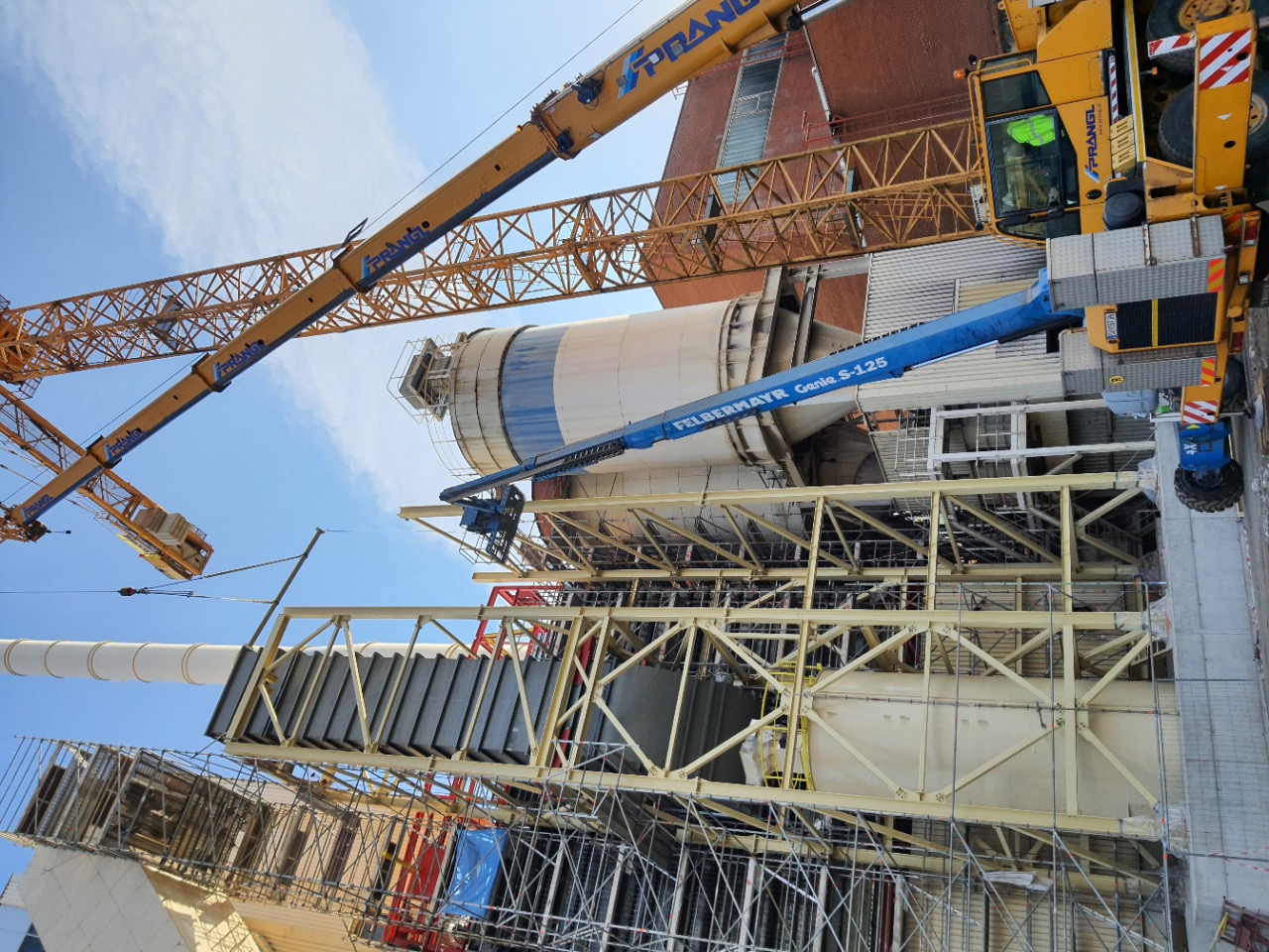 Wznoszenie konstrukcji stalowej hali przemysłowej z użyciem kilku dźwigów, widoczne elementy szkieletu i silosy w tle, niebieski podnośnik koszowy marki Felbermayr.