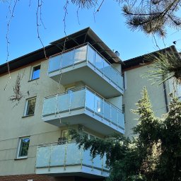 FRB AT Wróbel Sp. z o.o - Trzy balkony z balustradami ze szkła mlecznego na budynku wielorodzinnym, widok z dołu, częściowo przysłonięte gałęziami drzew iglastych, drabina oparta o ścianę na dole.