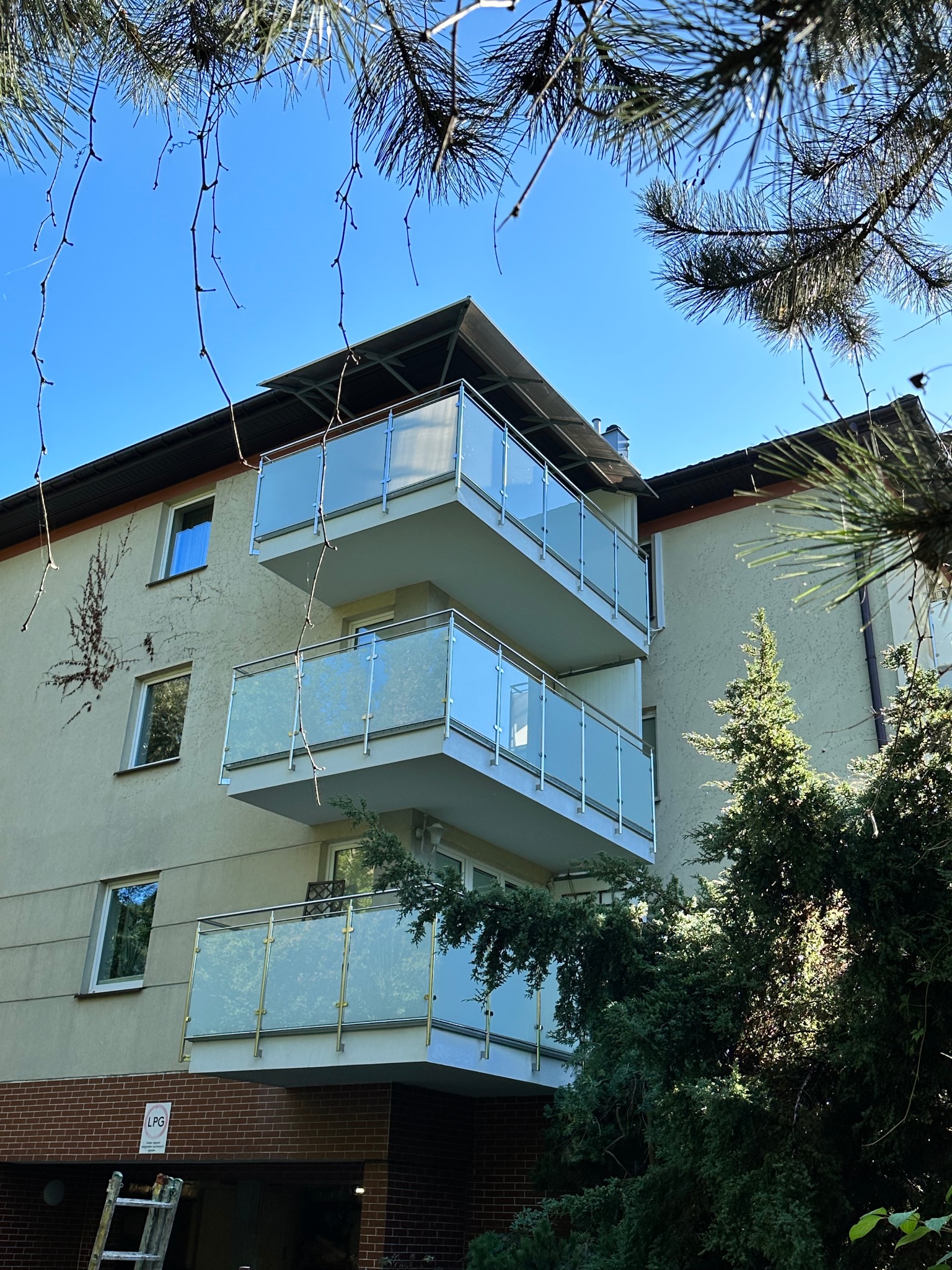 Trzy balkony z balustradami ze szkła mlecznego na budynku wielorodzinnym, widok z dołu, częściowo przysłonięte gałęziami drzew iglastych, drabina oparta o ścianę na dole.