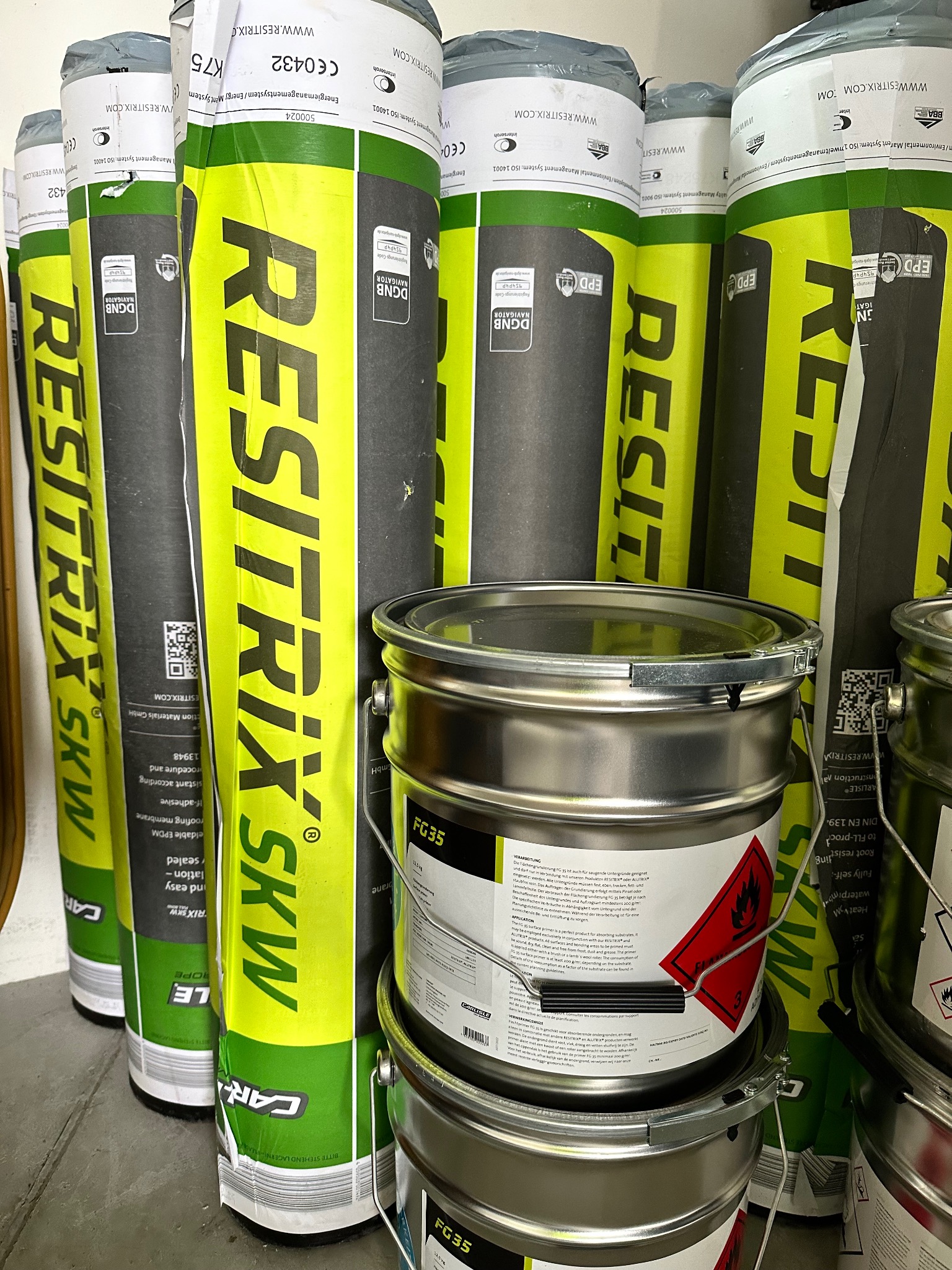 Stosy rolek membrany dachowej Resitrix SKW w zielono-czarnym opakowaniu, obok stoją wiadra z gruntem FG35, widoczne etykiety z informacjami technicznymi.