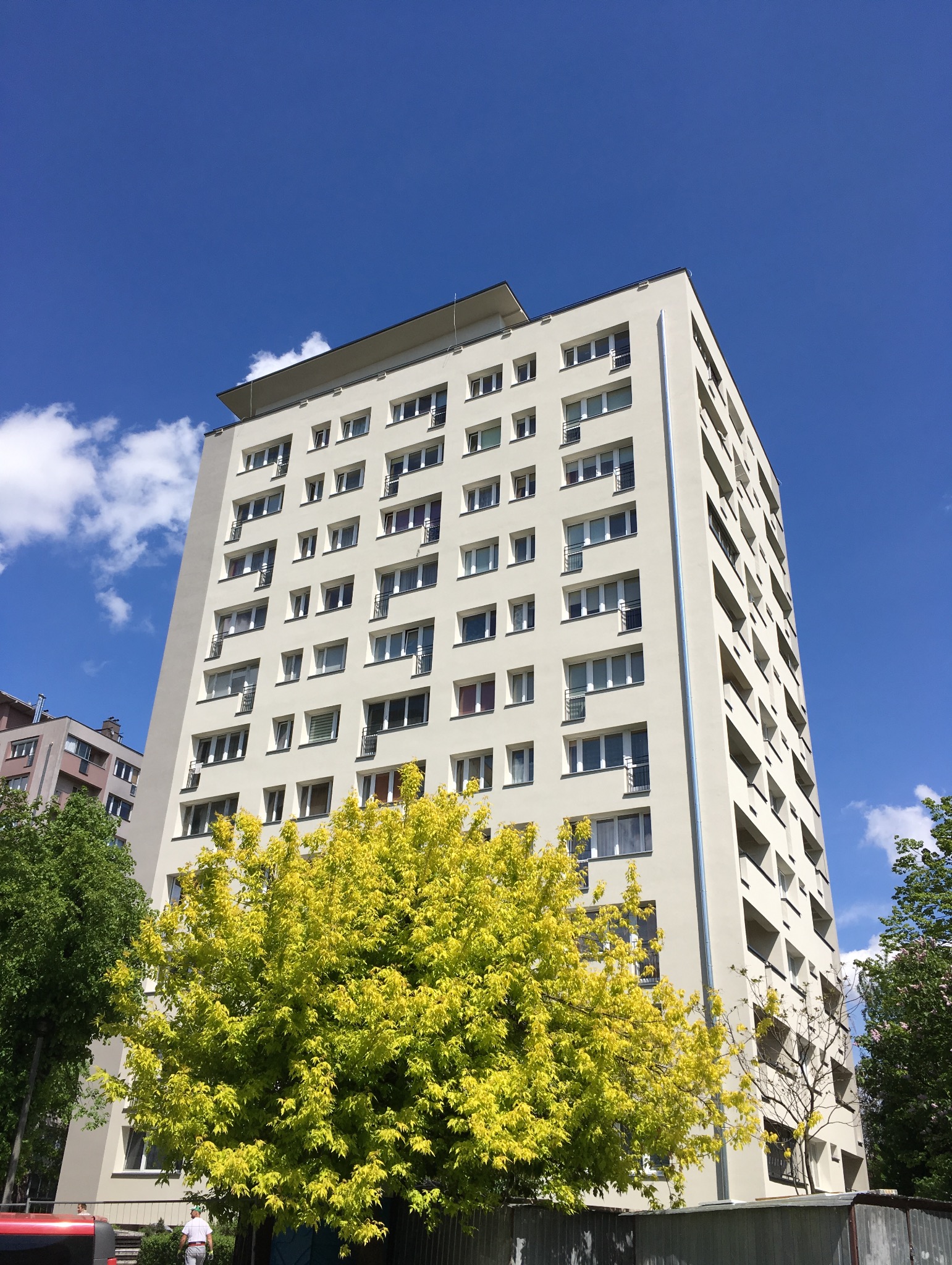 Wysoki budynek mieszkalny z jasną elewacją, widoczny z dołu, częściowo zasłonięty przez drzewo o żółtych liściach, na tle błękitnego nieba z chmurami.