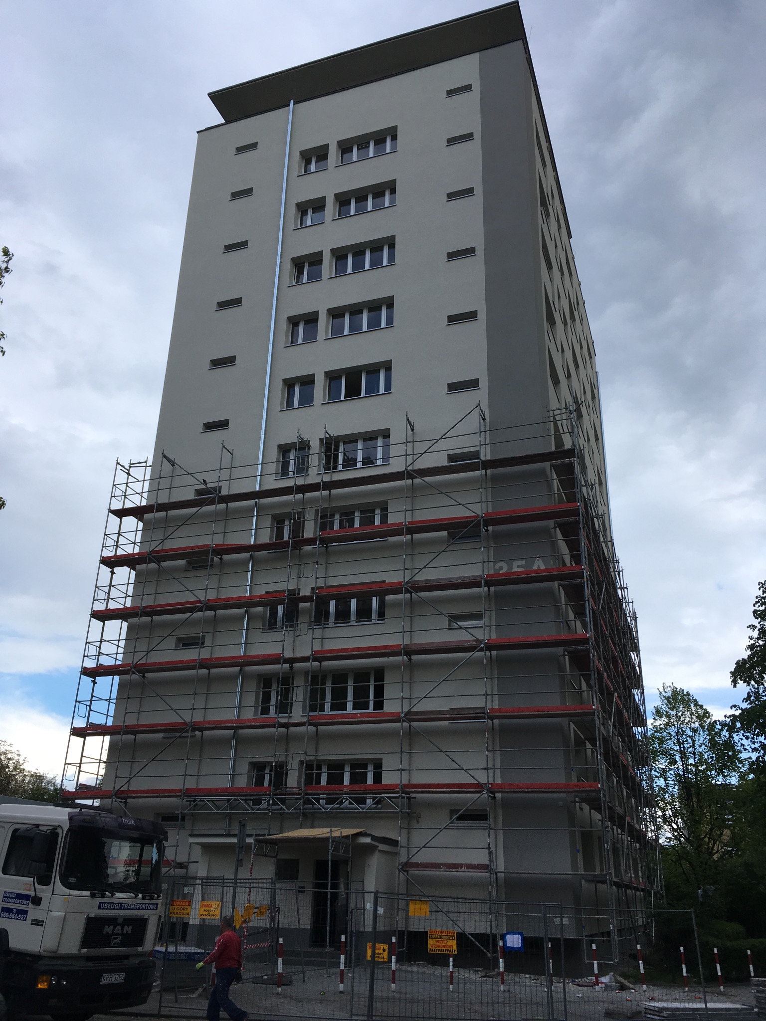 Wysoki budynek mieszkalny w trakcie prac elewacyjnych, otoczony rusztowaniami, widoczny fragment ciężarówki transportowej i ogrodzenie terenu budowy.
