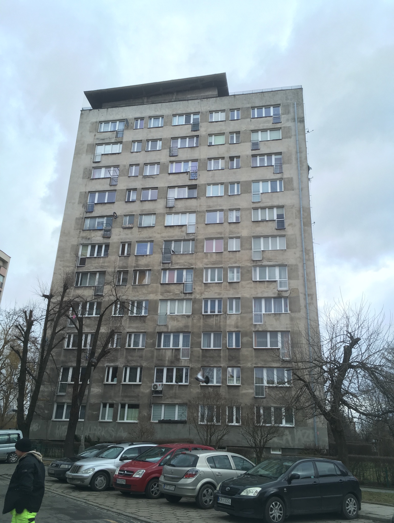 Wysoki, szary blok mieszkalny z balkonami, widok z dołu na pochmurnym niebie, zaparkowane samochody na pierwszym planie.