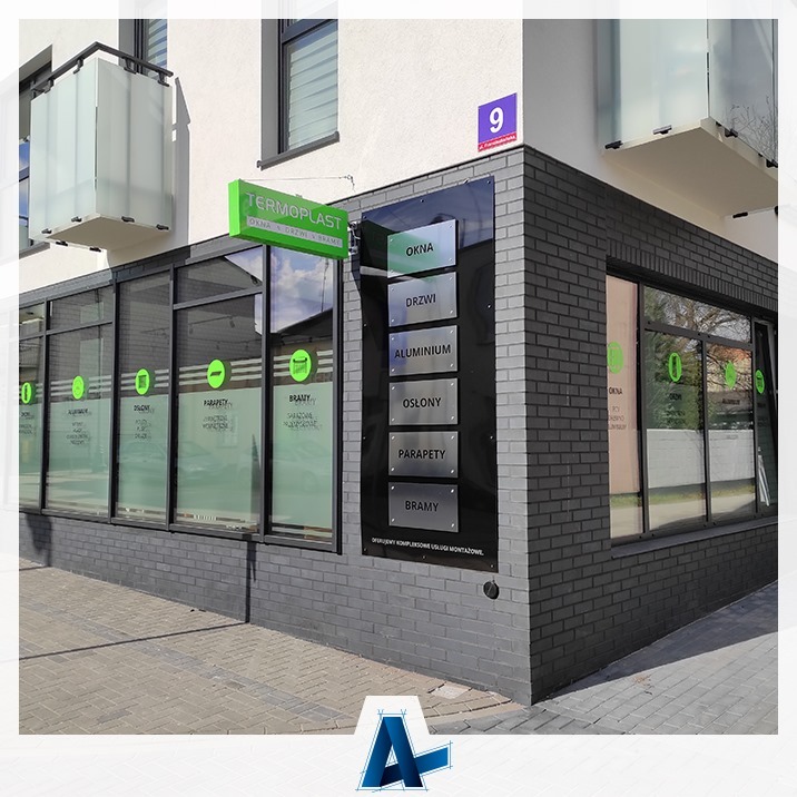 Fasada budynku firmy Termoplast oferującej okna, drzwi, bramy i inne elementy, widoczny szyld z nazwą firmy, tablica informacyjna z zakresem usług i witryny okienne z naklejonym logo firmy.