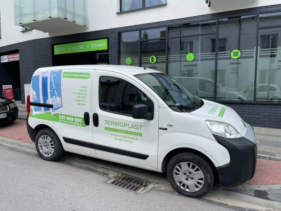 Biały samochód dostawczy firmy Termoplast z reklamą okien, drzwi i bram na tle budynku z oknami i szyldem reklamowym firmy.