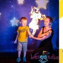 Kobieta w czarnej sukience prezentuje płonący rekwizyt w kształcie konia chłopcu w żółtej koszulce na tle dekoracji z gwiazdami, z widocznym logotypem firmy.