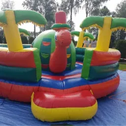 Kolorowy, dmuchany zamek w kształcie statku z palmami na trawie, rozłożony na niebieskiej plandece.