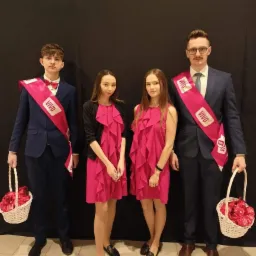 Grupa czterech osób: dwóch mężczyzn w garniturach z szarfami i koszykami czerwonych serduszek oraz dwie kobiety w różowych sukienkach, stojąca na tle czarnej ściany.