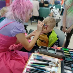 Animator z różowymi włosami maluje wzór na twarzy chłopca, używając pędzla i farb do twarzy. Na stole widoczne palety z farbami i pędzle.