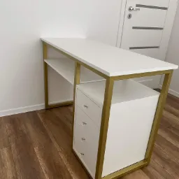 Białe biurko z trzema szufladami i złotą metalową ramą, na drewnianej podłodze, na tle białej ściany i białych drzwi.