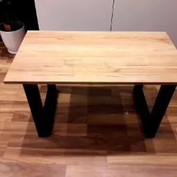 Blat drewniany o jasnym odcieniu drewna z delikatnym usłojeniem, wsparty na dwóch czarnych, trapezoidalnych nogach, stojący na podłodze imitującej drewno. W tle widoczna biała donica z rośliną...