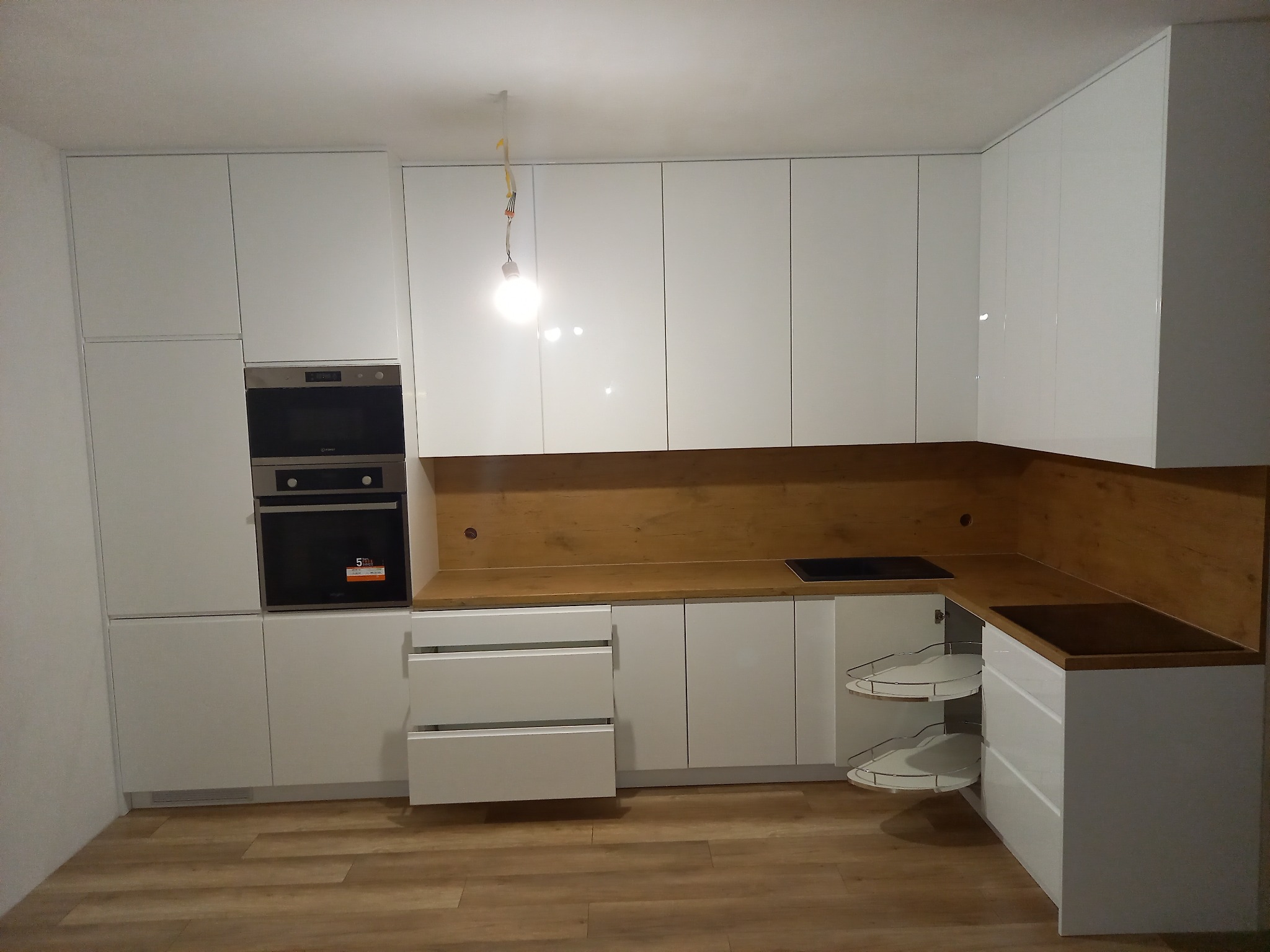 Nowoczesna kuchnia z białymi, lakierowanymi frontami, drewnianym blatem i narożną szafką z półkami obrotowymi; widoczny piekarnik w zabudowie.