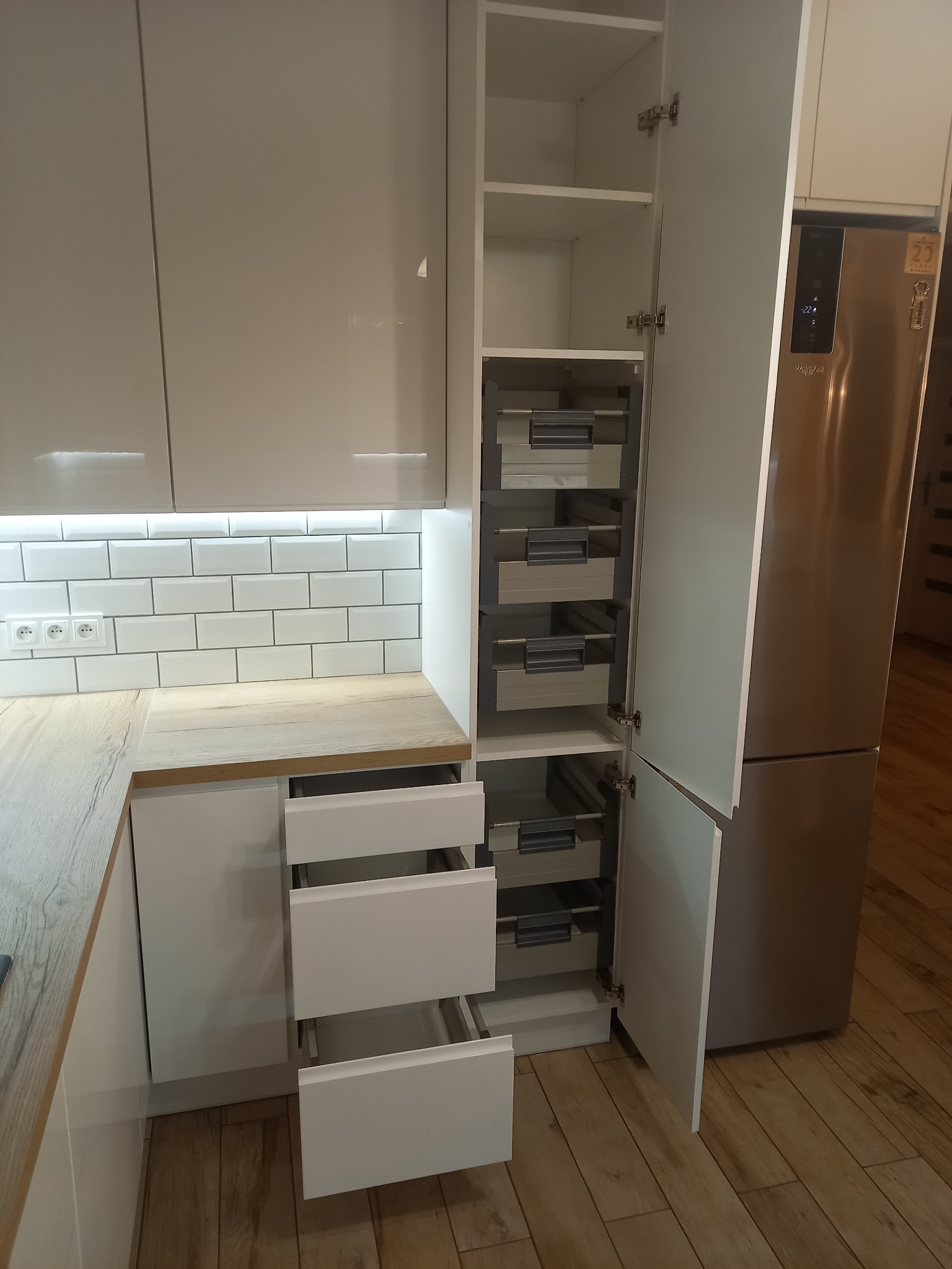 Nowoczesna kuchnia z białymi, lakierowanymi frontami, drewnianym blatem i otwartymi szufladami cargo oraz standardowymi, widoczna lodówka w zabudowie.