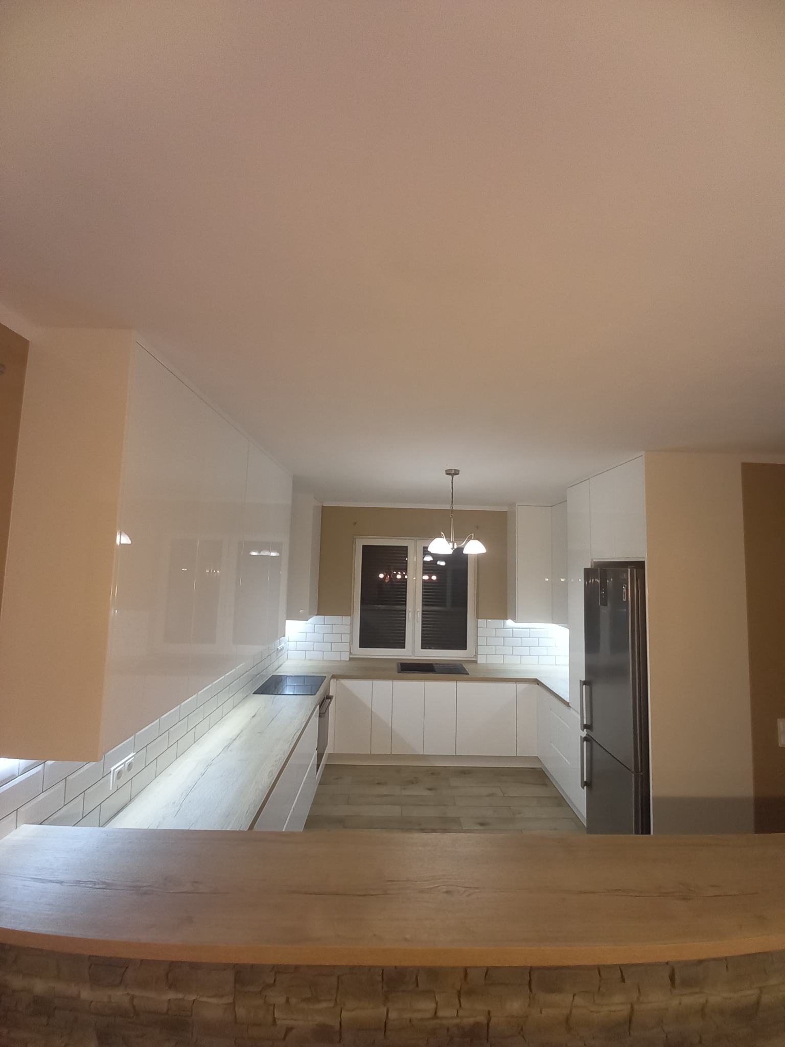 Nowoczesna kuchnia z białymi, lakierowanymi frontami, drewnianym blatem i barkiem wykończonym kamieniem dekoracyjnym, oświetlona lampą wiszącą i taśmami LED pod szafkami.