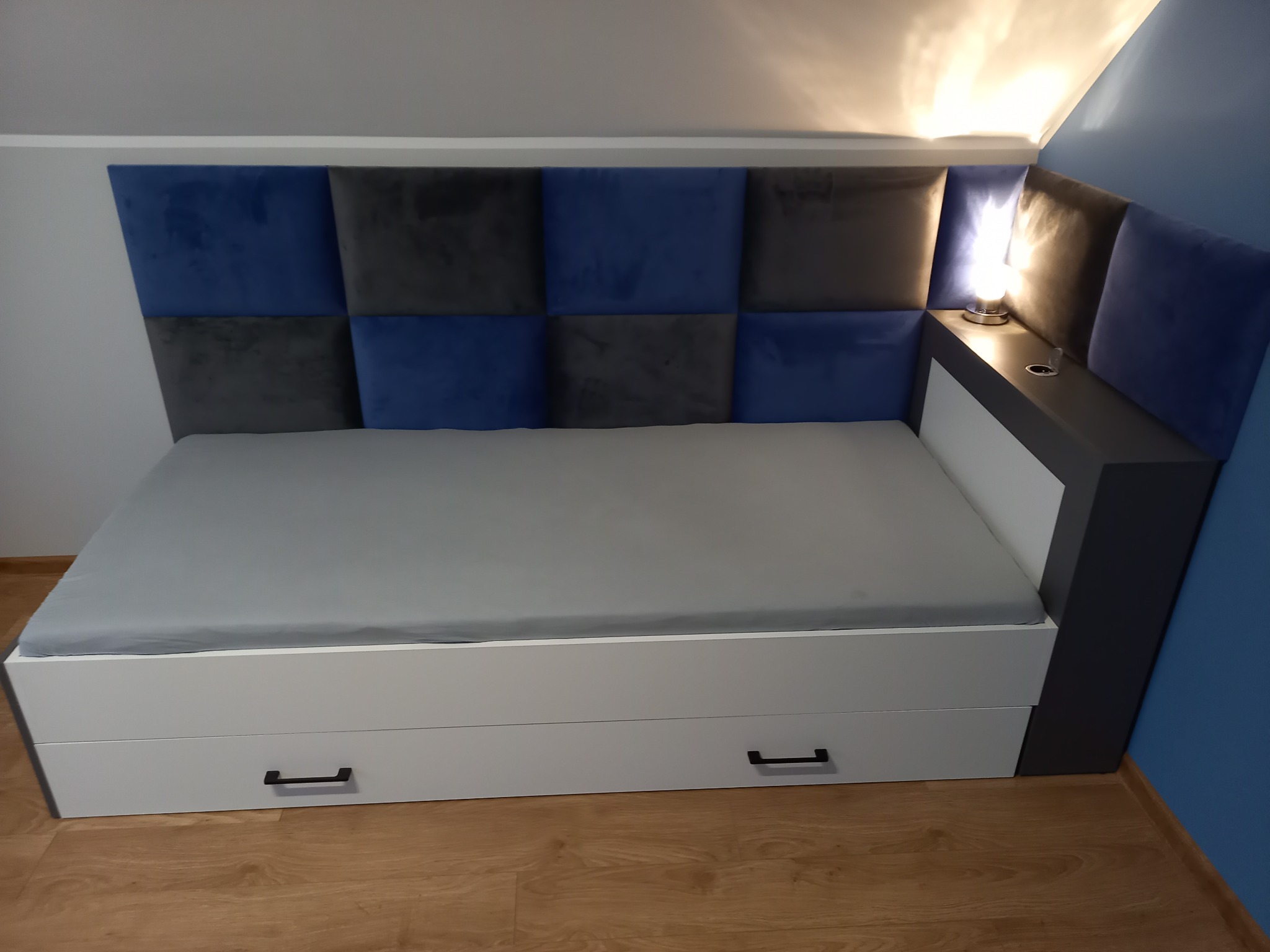 Łóżko z szufladami, tapicerowanym wezgłowiem w kratę z paneli w odcieniach szarości i granatu oraz szafką nocną z lampką.