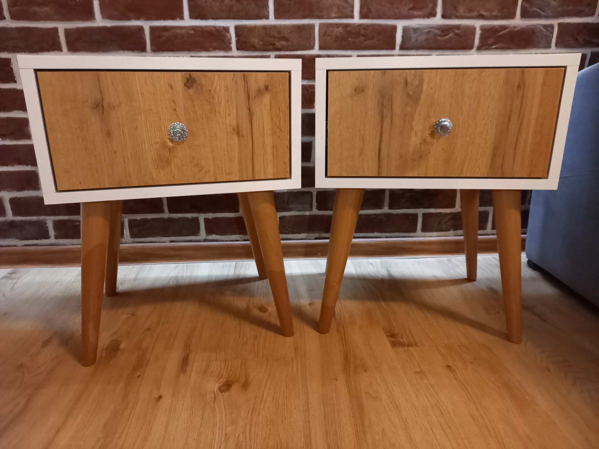 Dwie drewniane szafki nocne z szufladami i białymi ramami, ustawione na drewnianej podłodze na tle ściany z czerwonej cegły.