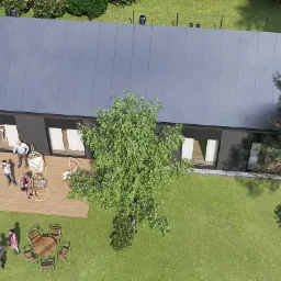 Wizualizacja z lotu ptaka nowoczesnego, parterowego domu z ciemną elewacją i szarym dachem na zadbanym trawniku, z widocznym tarasem, meblami ogrodowymi i kilkoma samochodami na podjeździe.