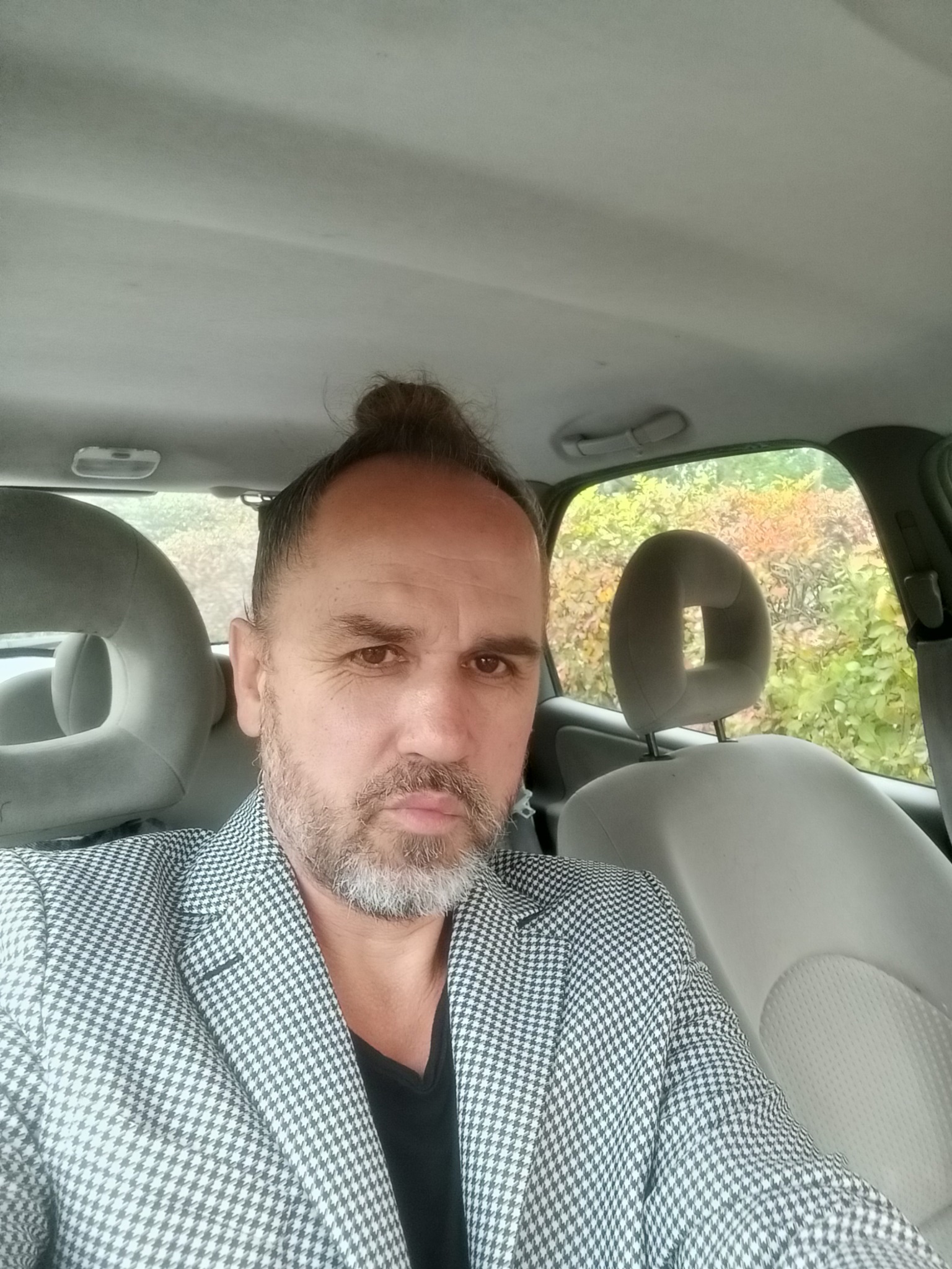 Mężczyzna z brodą i w marynarce w pepitkę siedzi w samochodzie, widok z przodu.