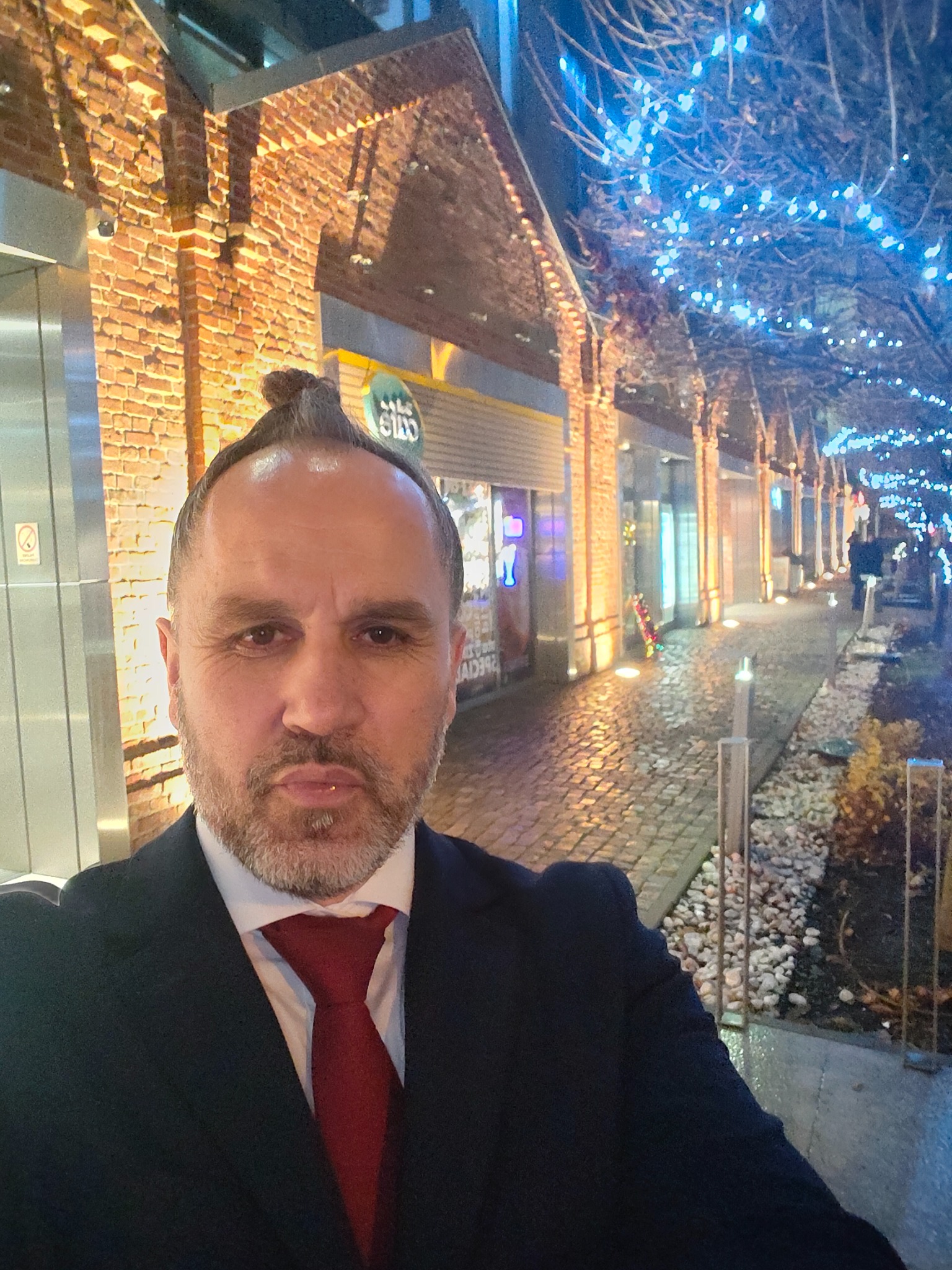 Portret mężczyzny w garniturze i czerwonym krawacie na tle oświetlonej ulicy z ceglanymi budynkami i drzewami z niebieskimi lampkami. Nietypowa fryzura.